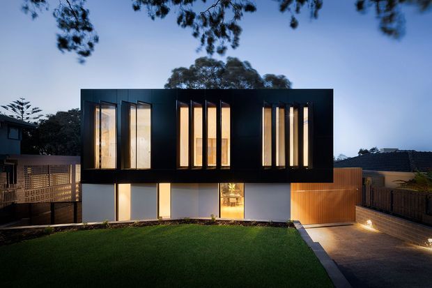 Twilight exterior view of a contemporary two-storey residence with a bold dark upper volume, tall narrow vertical windows, illuminated lower-level glazing, timber screening, and a paved driveway leading to the garage. The home sits in a landscaped suburban setting with a broad front lawn.