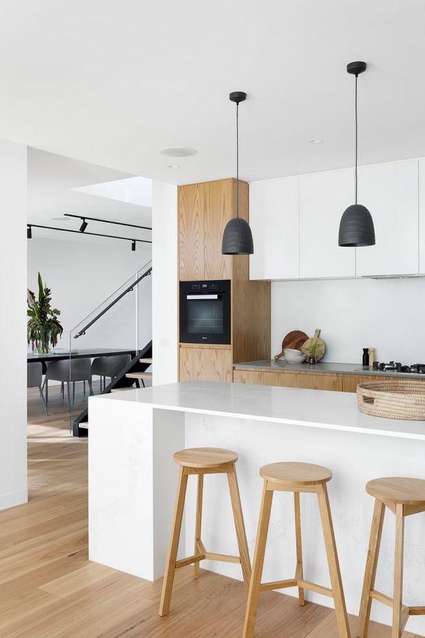 Bright open-plan kitchen with a large white waterfall island and integrated breakfast bar, pale timber stools, white full-height cabinetry, and a timber veneer oven housing. A dark stone or composite benchtop runs along the rear wall, with black pendant lights and track lighting overhead. Light timber flooring continues into the adjacent dining area and stairway, creating a clean, contemporary minimalist interior.
