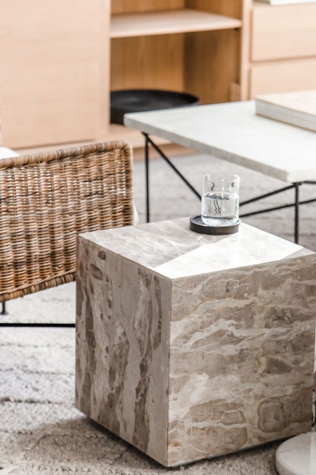 Close-up of a refined living room seating area featuring a sculptural beige stone coffee table, a pale stone-topped side table, a woven rattan chair, and light wood built-in cabinetry. The space has a warm neutral palette and minimalist contemporary styling.