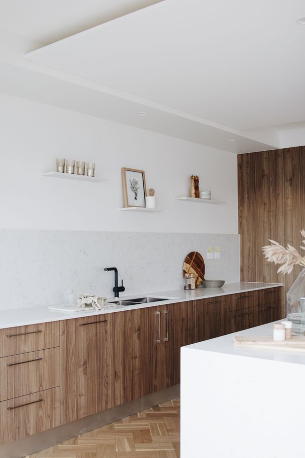 Bright contemporary kitchen with full-height and base cabinetry in richly grained dark timber, a pale stone benchtop and splashback, integrated double sink with matte black tap, open display shelves, and a white island in the foreground. Herringbone timber flooring and recessed ceiling lighting complete the refined, warm-toned interior.