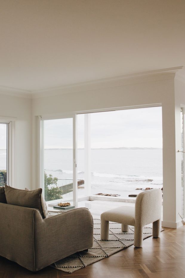 Bright, refined living room overlooking the sea through expansive glazed doors and windows. The room features a neutral upholstered armchair and sofa, a pale textured rug, herringbone timber flooring, white walls with decorative ceiling cornices, and abundant natural light.