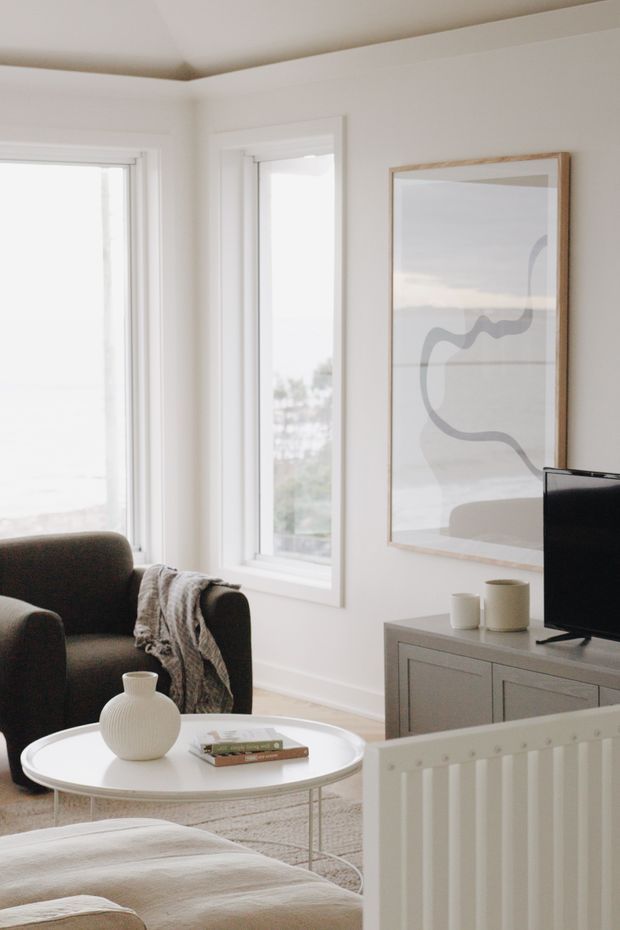 Bright, calm living room with white walls, tall windows, a dark upholstered armchair, round white coffee table, pale upholstered seating in the foreground, and a grey built-in-style media console. A large abstract framed artwork and neutral textiles add understated visual interest.