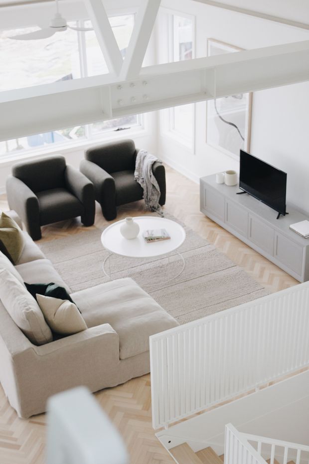 Bright, airy living room viewed from an elevated stair or landing position. The room features a large light upholstered sectional sofa, two dark lounge chairs, a white round coffee table, a pale textured rug, a low white media console with television, herringbone timber flooring, large windows, and prominent white exposed structural beams. A white balustrade borders the foreground.
