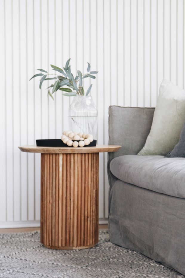 A close-up interior vignette featuring a fluted cylindrical timber side table beside a grey upholstered sofa. The table is styled with a black tray, pale wooden beads, and a textured white vase with leafy branches. A white vertical wall panelling backdrop and neutral patterned rug create a soft, refined palette.