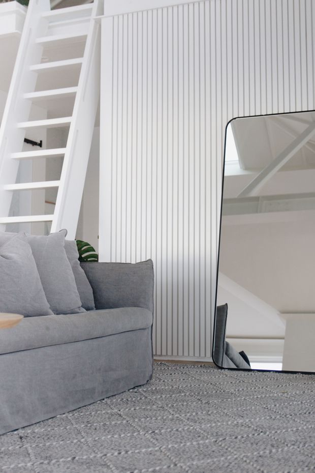 Bright, minimalist living room with a pale grey sofa, textured grey rug, white vertical wall panelling, and a large leaning black-framed mirror. A white ladder-style stair or loft access structure and exposed white beams are visible, creating an airy contemporary interior.
