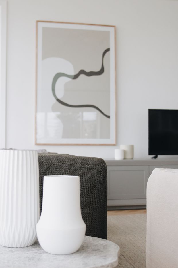 A bright, softly styled contemporary living room with a neutral palette. The scene features a dark textured armchair, a pale upholstered sofa, a low light-coloured built-in media console with television, a round stone or marble coffee table, and minimalist white ceramic vessels. A framed abstract artwork hangs on the white wall, with daylight reflected in the glossy surface.