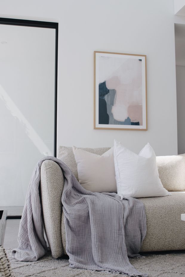 A bright, softly styled living room corner featuring a light beige textured sofa with white cushions and a draped pale grey throw. A large black-framed glazed opening or mirror is at left, with minimalist abstract artwork in a light timber frame above the sofa. The space has a restrained neutral palette and abundant natural light.