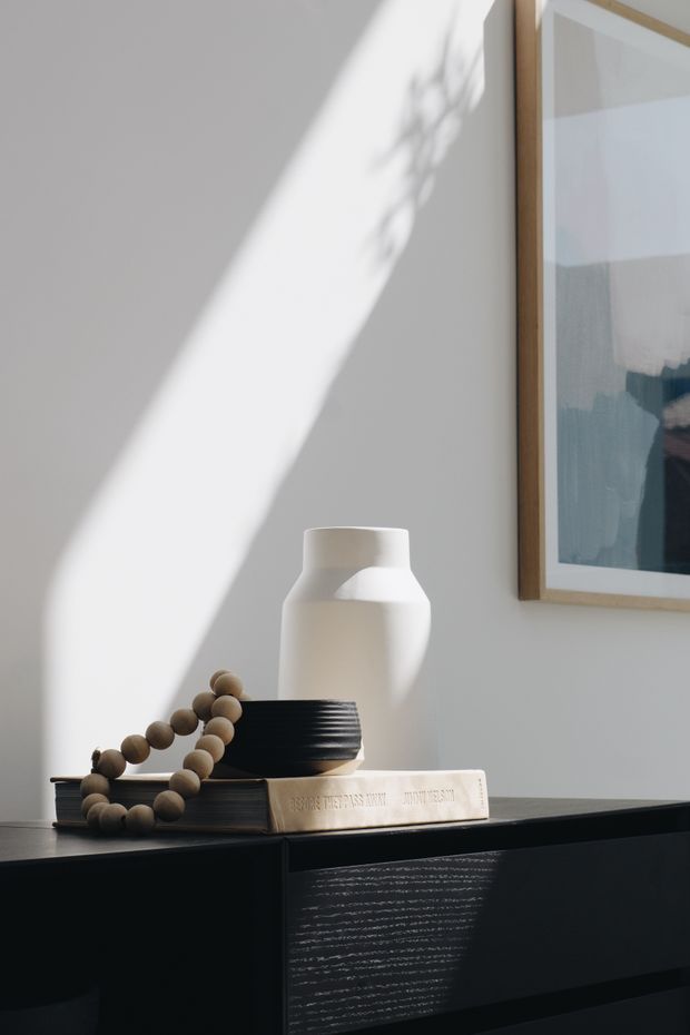 A styled interior vignette featuring a dark black built-in or freestanding console cabinet against a white wall. A white sculptural ceramic vase, stacked books, and a wooden bead garland sit on top, with framed abstract artwork partially visible above. Strong diagonal sunlight creates dramatic shadows across the wall and cabinetry.