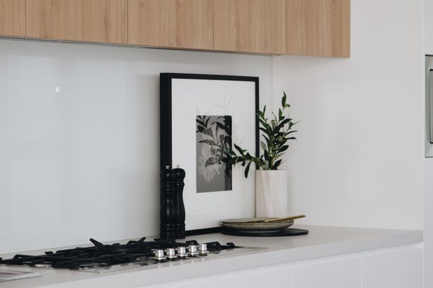 A clean contemporary kitchen vignette with a white stone benchtop, integrated gas cooktop and black fixtures. Light timber overhead cabinetry spans the upper wall, while a framed botanical print, black pepper mill, ceramic vase with leafy branches and shallow decorative bowl add restrained styling.
