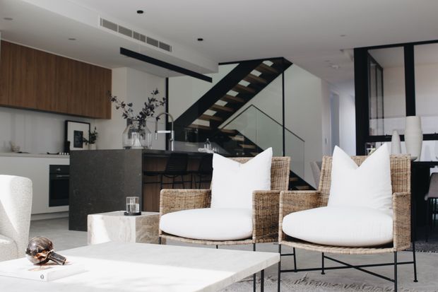 A refined open-plan living and kitchen interior with a dark stone island, timber cabinetry, woven lounge chairs, and a sculptural black-framed staircase behind a glass balustrade. The predominantly white and light-grey scheme is accented by warm timber and natural woven textures, creating a bright, contemporary space.