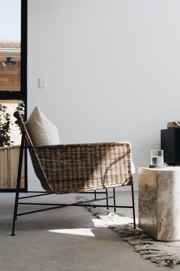 A sunlit modern living room corner featuring a woven rattan lounge chair with a dark metal frame, a pale cushion, a polished stone side table, and a glass of water. The room has white walls, a grey concrete-look floor, large black-framed glazing, and a neutral textured rug. The styling is restrained, natural, and contemporary.