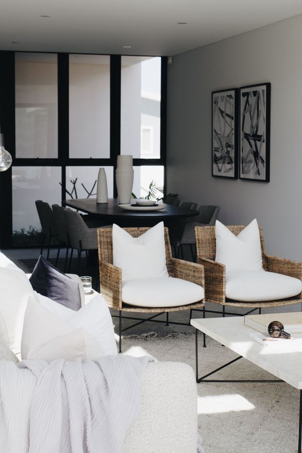 A bright, refined open-plan living and dining area. The foreground features a white upholstered sofa with textured throw and cushions, two woven rattan armchairs, and a minimalist white rectangular coffee table. Behind them is a dark dining table with upholstered chairs, sculptural ceramic vases, and abstract monochrome artwork. Floor-to-ceiling black-framed glazing and a neutral grey-white palette create a contemporary, airy interior.
