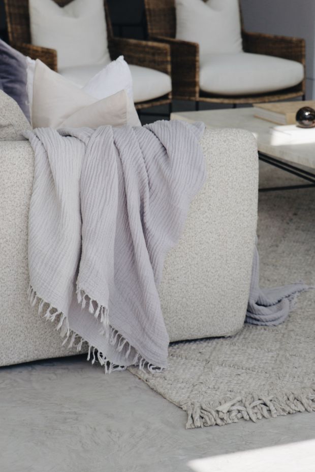 Close-up of a softly styled contemporary living room featuring a light textured upholstered sofa, pale lavender throw blanket with fringed edges, white cushions, a textured area rug, woven armchairs in the background, and a marble-top coffee table. The palette is light, neutral, and understated with natural woven accents.