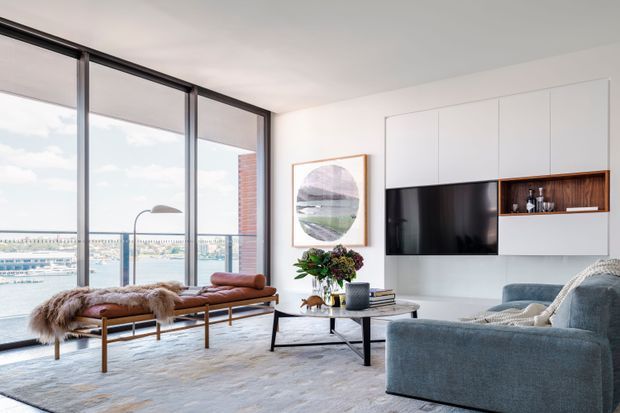 Bright contemporary living room with floor-to-ceiling glazing overlooking a waterfront or marina. The space features a low built-in white media wall with timber display shelving, a large television, a blue-grey sofa, a leather-and-timber bench with fur throws, a marble or stone coffee table, and restrained decorative styling.