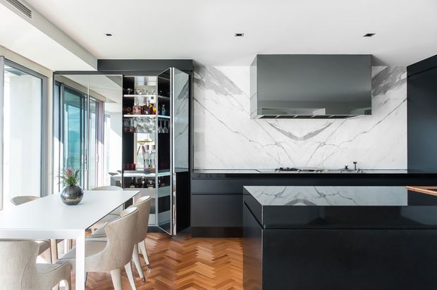 Contemporary open-plan kitchen and dining area with dark matte cabinetry, a large white marble-look splashback and matching island face, integrated rangehood, glass-fronted drinks cabinet, and a light dining table with upholstered chairs. Floor-to-ceiling glazing brings in abundant daylight, while the warm herringbone timber floor contrasts with the monochrome kitchen palette.