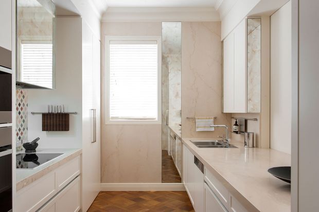 Bright, narrow galley-style kitchen with white shaker cabinetry, pale stone benchtops, marble-look wall finishes, stainless steel sink and tapware, integrated appliances, and a central window with white plantation shutters. Herringbone timber flooring adds warmth, while a small patterned tile splashback and mirrored surfaces provide decorative detail.