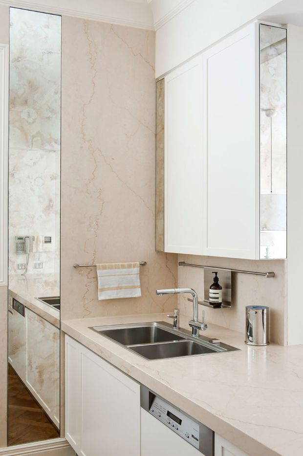 A bright, refined bathroom vanity area with a double stainless-steel sink set into a pale stone countertop, white shaker-style cabinetry, mirrored side panels, chrome fixtures, and a large beige-veined stone backsplash and wall surface. A towel rail, soap dispenser, dishwasher-like under-bench appliance, and reflected cabinetry are visible. The space has a clean, elegant, traditional-influenced fit-out.