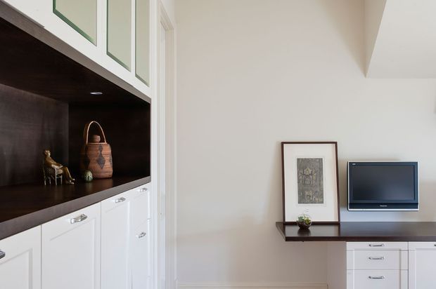 A refined built-in living room display and media unit. White shaker-style cabinetry with chrome pulls surrounds a dark timber open shelving niche containing decorative objects. A slim dark timber console supports framed artwork and a wall-mounted television, creating a clean, understated traditional interior.