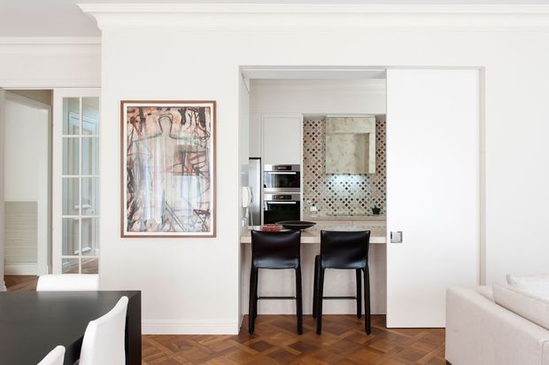 Bright, refined interior view showing a compact kitchen opening onto a living or dining area. The kitchen has dark lower cabinetry, integrated stainless-steel appliances, a patterned mosaic tile splashback, and a small peninsula with two black bar stools. White walls, detailed cornice moulding, glazed internal doors, and a herringbone timber floor create a classic architectural backdrop, while a large framed artwork and partial upholstered seating are visible in the adjacent space.