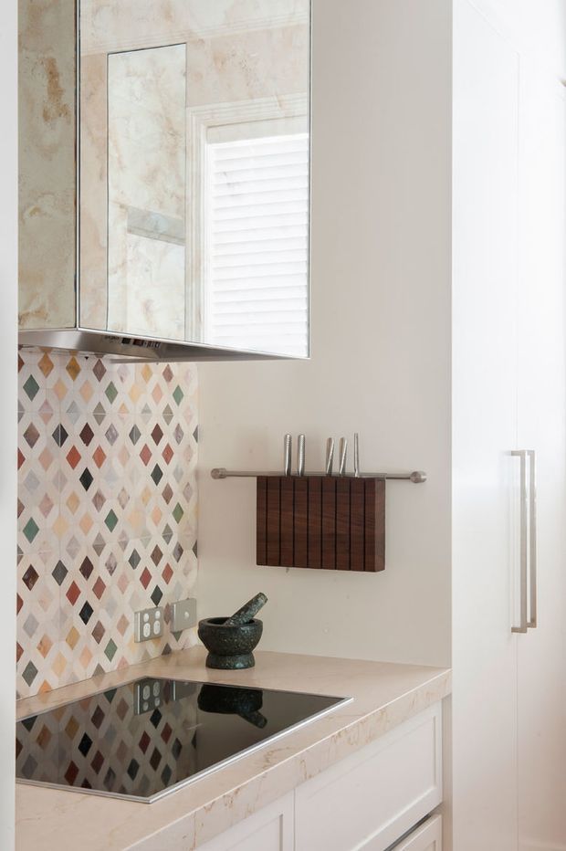 A bright, compact kitchen featuring white built-in cabinetry, a pale stone-look benchtop, black glass induction cooktop, mirrored overhead cabinetry, and a colourful geometric diamond-pattern tiled splashback. A wall-mounted wooden knife rack and dark stone mortar and pestle add functional detail.