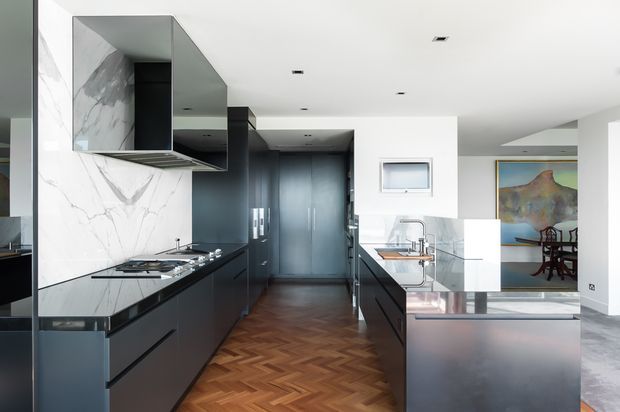 Contemporary galley-style kitchen with dark matte cabinetry, black stone benchtops, a dramatic white marble splashback, integrated stainless steel appliances, and warm herringbone timber flooring. A reflective island or peninsula separates the kitchen from an adjacent dining or living area, with artwork visible beyond.