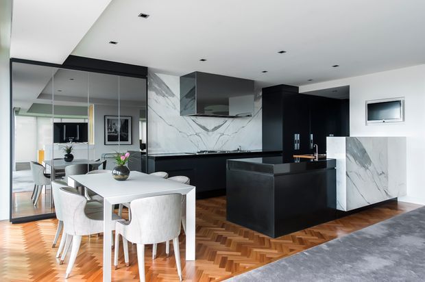A contemporary open-plan kitchen and dining area with a dramatic black-and-white palette. The kitchen features matte black cabinetry, a large black island, white marble-look splashback and side panels, integrated appliances, and a dark stone benchtop. A white dining table with upholstered chairs sits on a warm herringbone timber floor, while full-height glazing and mirrored panels expand the space visually.