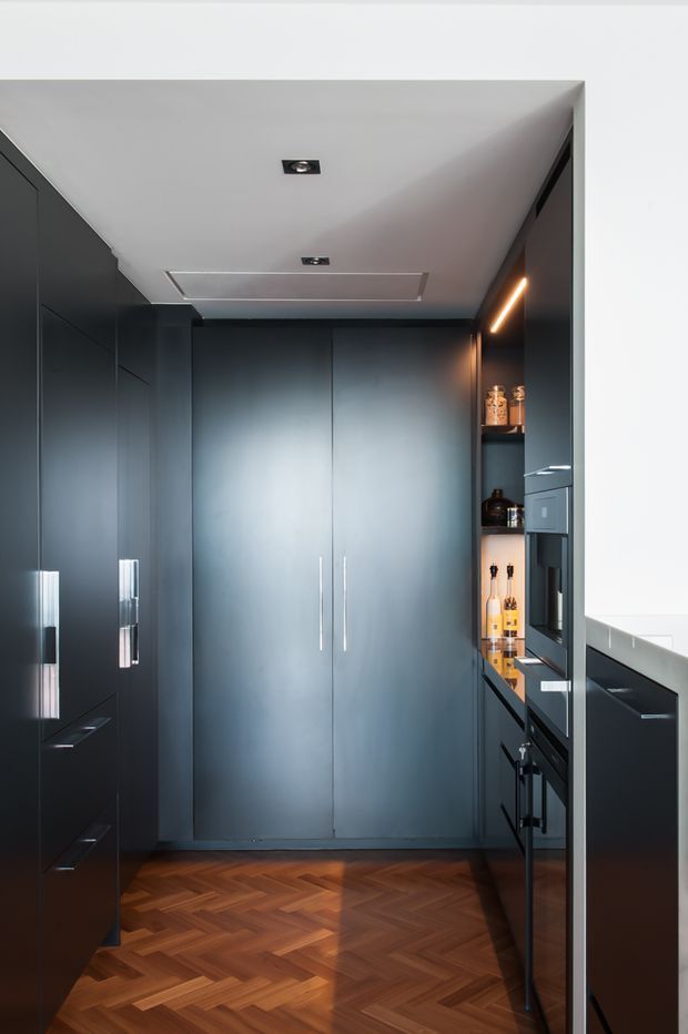 A narrow, contemporary galley kitchen with full-height dark blue-grey cabinetry, integrated appliances, open shelving with bottles and jars, and slim warm LED lighting. The room has a white ceiling with recessed downlights and a rich timber herringbone parquet floor. Minimal brushed-metal cabinet handles and a compact dark benchtop create a sleek, moody, high-contrast interior.