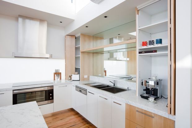 Bright contemporary kitchen with white cabinetry, pale timber joinery, a marble-look stone benchtop, stainless-steel appliances and a large mirrored splashback. The compact space includes a peninsula or island, integrated oven and cooktop, double sink, open shelving, and a built-in coffee station. Skylight-like overhead glazing and recessed lighting contribute to the airy feel.