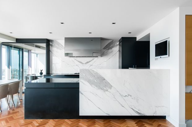 A bright contemporary open-plan kitchen with a large white marble-look island and waterfall benchtop, dark charcoal cabinetry, integrated stainless-steel rangehood, and a matching marble splashback. Floor-to-ceiling glazing opens toward a dining or living area, while warm herringbone timber flooring adds contrast.