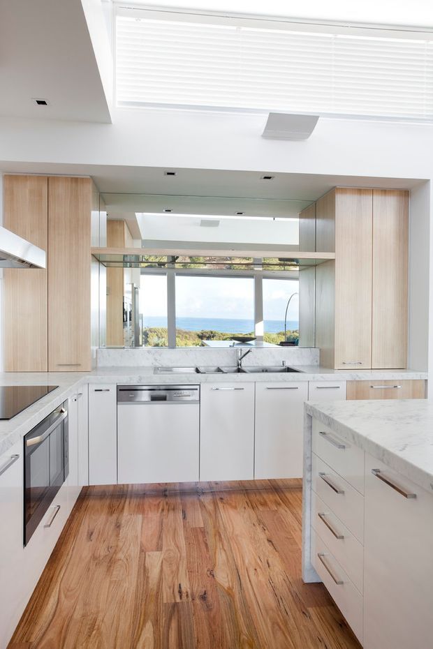 Bright contemporary kitchen with white cabinetry, pale timber overhead cabinetry, marble-look stone benchtops and splashback, stainless-steel appliances, and a central island. A large skylight and mirrored rear wall increase the sense of openness, while full-height glazing provides a coastal ocean view.