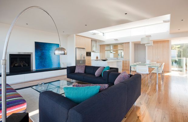 Bright open-plan contemporary living space with dark navy sectional sofas, a glass coffee table, a linear fireplace and blue artwork. The living room flows into a light timber kitchen and dining area, with extensive glazing and a skylit ceiling creating an airy atmosphere. A sculptural arched floor lamp and colourful cushions add contrast.