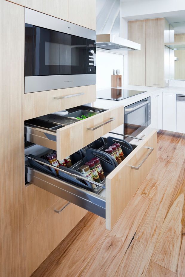 Bright contemporary kitchen featuring tall light timber cabinetry, integrated black-and-silver appliances, extensive pull-out pantry drawers, a white island with black glass cooktop, stainless steel handles, and warm timber flooring. The image emphasizes practical storage and kitchen joinery.