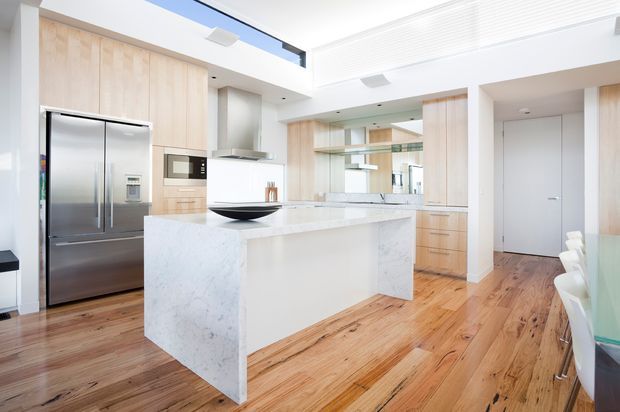 Bright, open-plan contemporary kitchen with a large white stone waterfall island, extensive light timber cabinetry, stainless-steel appliances, timber flooring, and high ceilings with clerestory glazing. A dining area and hallway are visible beyond the kitchen.