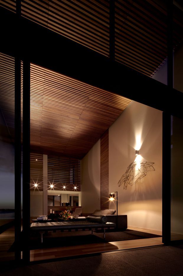 A dramatic, warmly lit contemporary living room viewed through dark-framed glazing. The space features a very high timber slatted ceiling and matching slatted screening, tall white walls, a low dark leather sofa, black coffee table, sculptural wall art, and layered architectural lighting. The composition emphasises the double-height interior and strong linear timber detailing.