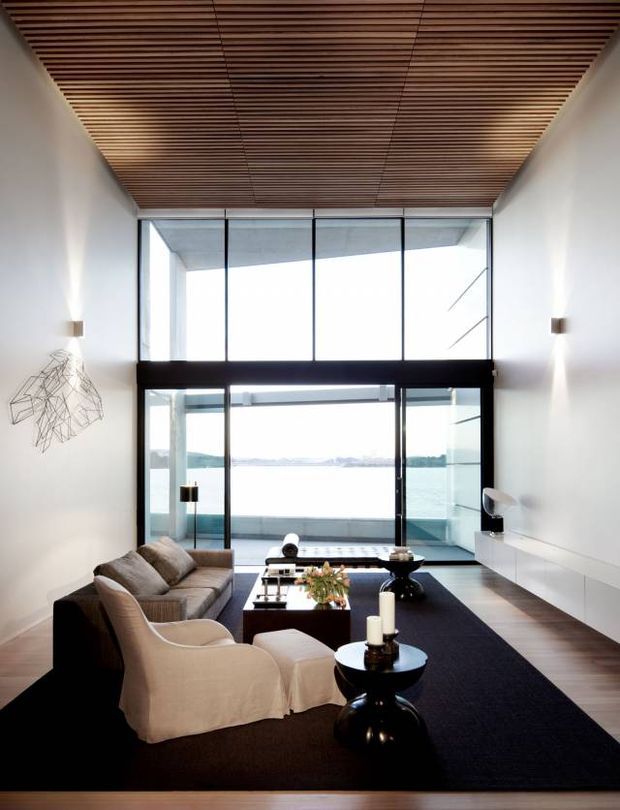 A refined double-height living room with expansive floor-to-ceiling glazing overlooking a body of water. The room features a warm timber slatted ceiling, white walls, dark-framed glass doors and windows, a large dark rug, a neutral sectional sofa and armchairs, sculptural side tables, and minimalist built-in white cabinetry. The composition is bright and spacious with a strong indoor-outdoor connection.