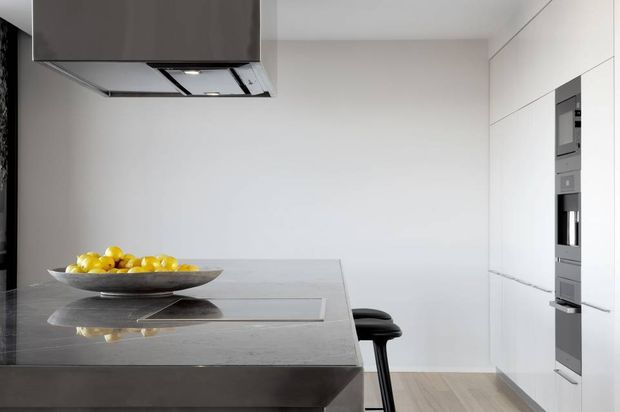 Minimal contemporary kitchen with a large dark grey stone island and integrated induction cooktop, paired with black bar stools. Full-height white cabinetry houses built-in stainless-steel appliances along the right wall. The space has plain white walls, light timber flooring, clean linear detailing, and a restrained monochromatic palette.