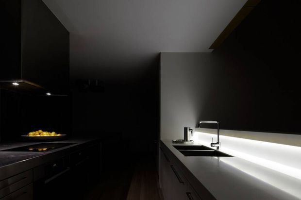 A dark, dramatic contemporary kitchen viewed along a long run of cabinetry and benchtop. The foreground includes a black cooking zone with integrated appliances and a shallow bowl of lemons, while the brighter right-hand side features a stainless-steel sink, minimalist chrome mixer tap, pale benchtop, and a continuous illuminated splashback or under-cabinet light. Flat-panel cabinetry, strong linear geometry, and concealed lighting create a moody, high-contrast atmosphere.