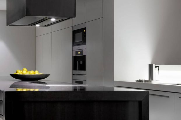 A sleek contemporary kitchen dominated by dark grey full-height cabinetry, integrated black appliances, and a large dark island with a reflective stone benchtop. A sculptural black fruit bowl provides a small yellow accent, while a bright window-side work area and stainless-steel tap add contrast. The space has clean, handleless lines and a restrained monochrome palette.