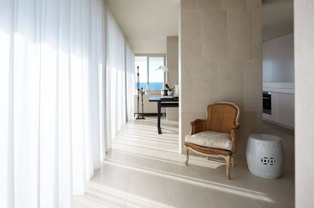 A bright, refined interior corridor or living area with full-height sheer curtains casting linear shadows across pale tiled flooring. A textured stone or porcelain-clad column divides the space, with a classic upholstered armchair and small white side table in the foreground. A minimalist desk and stool sit near large glazing at the far end, with a partial contemporary kitchen visible to the right and blue water or sky beyond the windows.