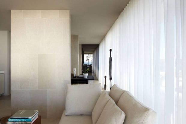 A bright, refined living room with a long cream sectional sofa positioned beside a full-height wall of sheer white curtains. The room features pale large-format stone or porcelain wall cladding, a neutral smooth ceiling, minimalist dark console joinery, and slender decorative floor lamps. A reflective opening at the rear extends the view through the space. The overall composition is calm, airy, and understated.