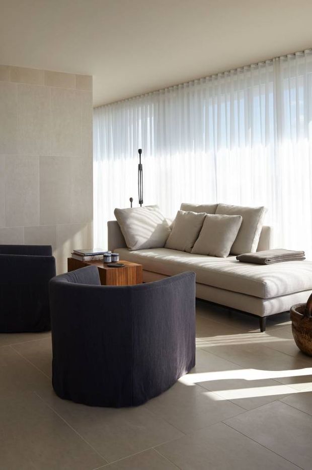 A bright, refined living room with a low cream sectional sofa, dark rounded lounge chairs, and a compact timber coffee table. The room has large curved glazing covered by sheer white curtains, a pale tiled feature wall, matching pale floor tiles, and strong natural sunlight creating long shadows. The styling is understated and contemporary with soft neutral upholstery and sculptural furniture.