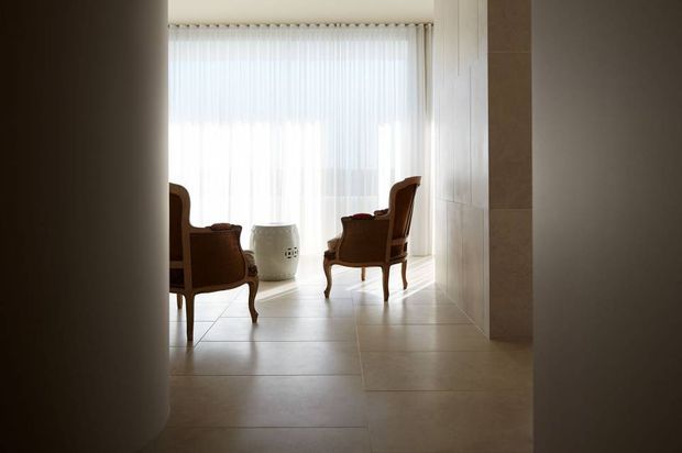 A softly lit, minimalist interior sitting area viewed through a narrow opening. Two classic upholstered wooden armchairs face inward around a small light-colored cylindrical side table, set against full-height sheer curtains and large pale floor tiles. A substantial pale stone or tiled wall plane frames the right side, while curved dark wall edges create a quiet, gallery-like composition.