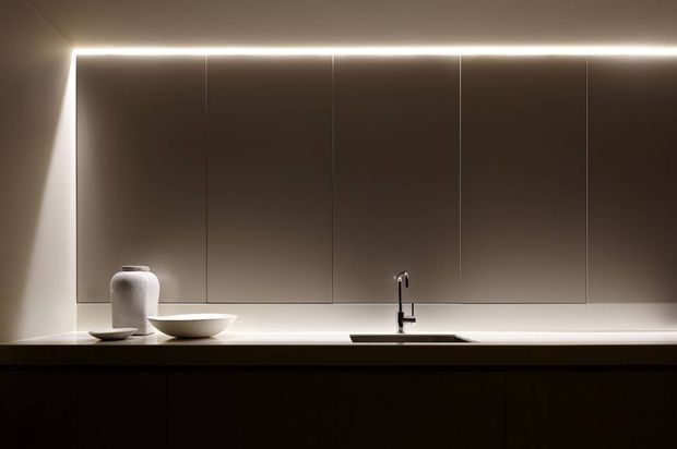 A minimalist contemporary kitchen presented in a dark, moody composition. The image shows a long white benchtop with an undermount sink and slim black tap, handleless dark taupe cabinetry extending to the ceiling, and integrated linear lighting along the upper edge. Simple white ceramic vessels sit on the counter, creating a restrained and refined contrast against the dark joinery.
