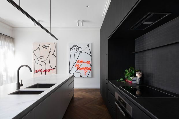 Contemporary kitchen with a large white stone island, integrated black sink and curved tap, dark floor-to-ceiling cabinetry, black tiled splashback, built-in appliances, and pendant lighting. White walls with decorative moulding and expressive framed artwork contrast with the dark joinery. The floor appears to be laid in a dark herringbone timber pattern.