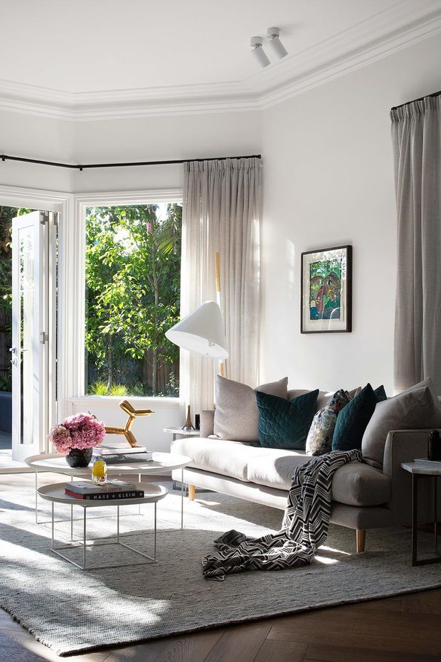 Bright, refined living room with white walls, decorative crown moulding, tall windows and an open glazed door overlooking greenery. A light upholstered sofa is styled with grey, teal and patterned cushions, accompanied by a sculptural floor lamp, round nesting coffee tables, artwork and a large neutral rug over dark timber flooring.