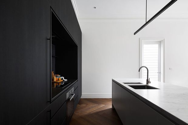 Contemporary kitchen interior featuring a large dark island with a pale stone benchtop, black full-height cabinetry and integrated appliances, white walls with decorative ceiling detailing, a black linear pendant light, and a herringbone timber floor. A window with white shutters provides natural light.