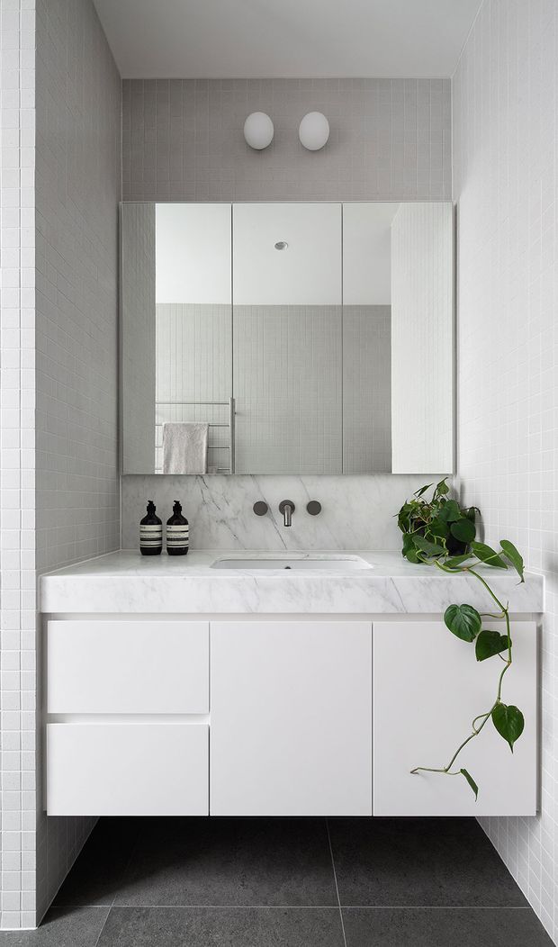 A compact, contemporary bathroom vanity with a wall-mounted white cabinet, white marble benchtop and integrated basin, brushed metal wall-mounted tapware, large mirrored medicine cabinet, white small-format wall tiles, and dark grey floor tiles. A trailing potted plant adds a natural accent.