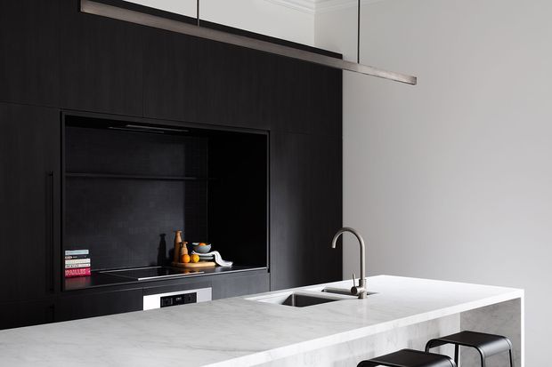 A refined contemporary kitchen dominated by a dark timber-toned full-height wall of cabinetry with an integrated black display niche and appliances. A large pale stone island with an inset sink and curved stainless-steel tap occupies the foreground, paired with dark stools. A slim linear pendant hangs above the island, creating a restrained, high-contrast monochromatic composition.
