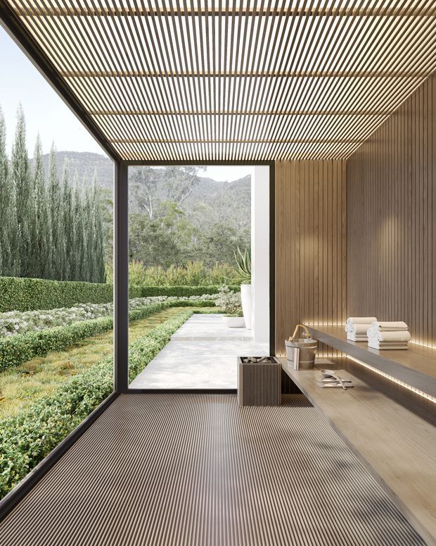 A refined, minimalist bathroom or spa-like bathing space with extensive floor-to-ceiling glazing opening toward a landscaped garden and mountain setting. The room features warm vertical timber wall cladding, a slatted timber ceiling and ribbed dark flooring, with a long built-in timber vanity or bench, integrated linear lighting, towels, a basin and a small waste bin. The open, sheltered design creates a strong connection to nature.
