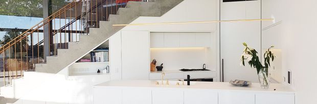 Bright contemporary open-plan kitchen beneath a sculptural concrete staircase. The kitchen features extensive white handleless cabinetry, a long white island with an integrated black tap and sink, a recessed illuminated display niche, and a dark benchtop cooking zone. Full-height glazing and a timber-and-black stair balustrade bring natural light into the space.